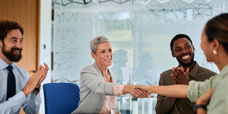 Four professionals sit at a conference table; a woman with short gray hair shakes hands with another woman, while two colleagues smile and applaud. Laptops and papers are on the table in a modern office setting.