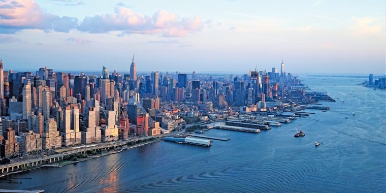 Aerial view of Manhattan, New York City at sunset, showing skyscrapers, waterfront docks, and boats moving on the Hudson River under a partly cloudy sky.