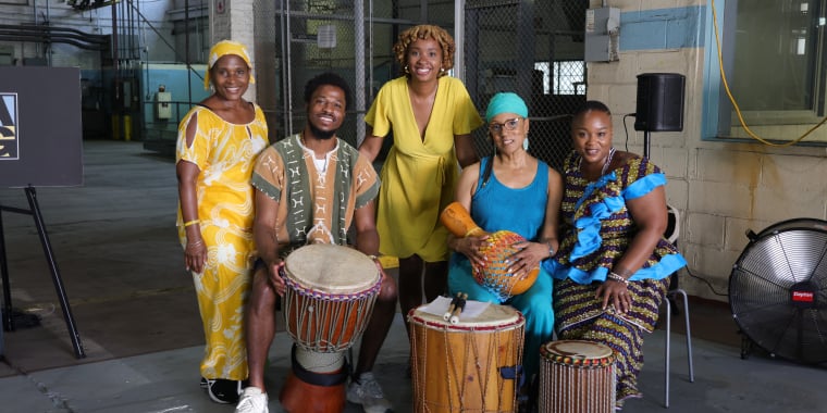 Five people in colorful, traditional African attire pose indoors with various drums and percussion instruments, smiling at the camera. The setting is an industrial-style room with exposed pipes and a large fan.