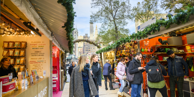 People browse festive Union Square Holiday Market stands decorated with greenery and lights. Vendors display snacks, gifts, and holiday items. Trees and tall buildings are visible in the background under a bright sky.