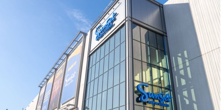 Image of Sunset Pier 94 Studios shows a modern building with large glass windows displays blue Sunset Pier 94 Studios sign. Three colorful billboards for the building are mounted on the side. The sky is clear and blue.