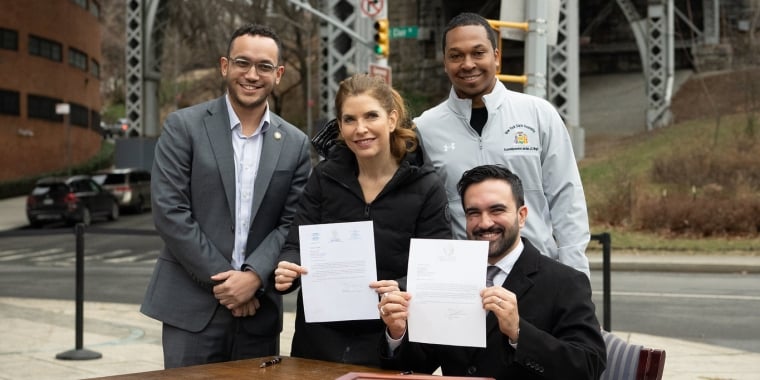 New York City Mayor Zohran Mamdani Mayor Mamdani and New York City Council Speaker Julie Menin holding up signed final approval documents for New York City Department of Transportation to install a new public bathroom later this year. Two men stand behind them. A road, sidewalk, and metal bridge structure are in the background. Image courtesy of Michael Appleton/Mayoral Photography Office.
