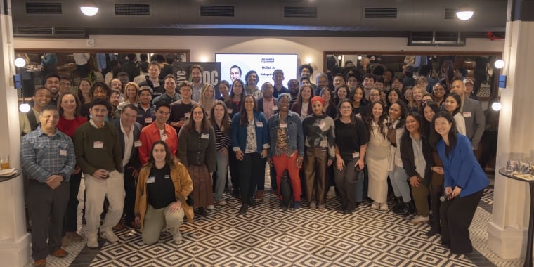 2026 Founder Fellowship cohort pose and smile for a group photo in a well-lit indoor space with patterned floors, mirrors, and a screen in the background. Most of them are wearing name tags.