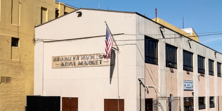A beige two-story building with an American flag and a sign reading “Brooklyn Wholesale Meat Market” on the front, located on a street corner with yellow-painted curbs.