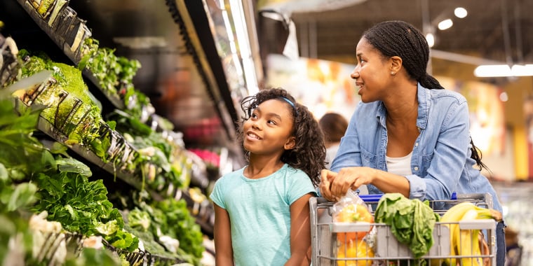 A woman and a young girl smile while shopping for fresh produce in a grocery store. The shopping cart is filled with fruits, vegetables, and groceries, and they are standing near the leafy greens section.