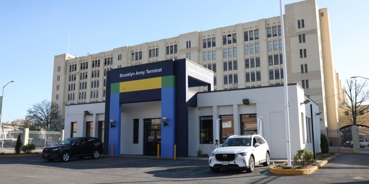 A small building with a colorful sign reading Brooklyn Army Terminal stands in front of a larger beige multi-story structure. Two cars are parked outside under a clear blue sky.