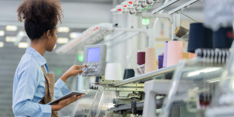 A woman wearing a blue shirt and apron operates a control panel in a modern textile factory, holding a digital tablet beside spools of colorful thread and automated machinery.