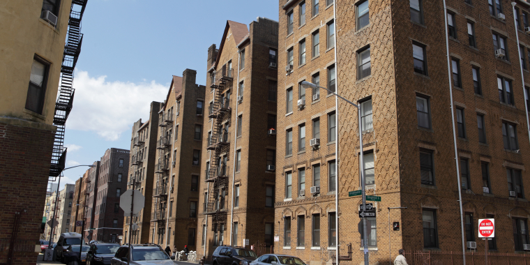 A street corner in Brighton Beach, Brooklyn with parked cars and tall brown brick apartment buildings. A person with a cane is walking on the sidewalk under a partly cloudy sky.