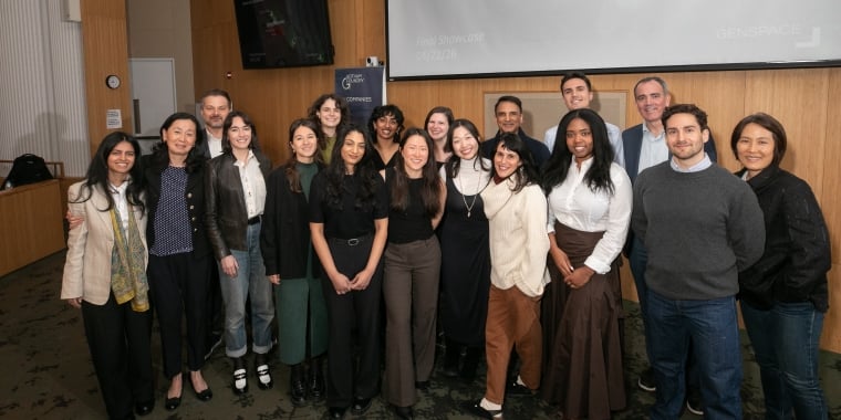 18 winners of Gotham Foundry Pitch Competition stand together and smile for a group photo in a conference room, with a projector screen and television visible in the background. Credit: Timothy Lee Photographers.