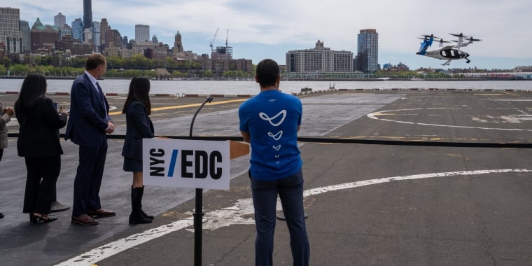 A small group of people stand near a NYC/EDC sign at Downtown Heliport, watching an electric vertical takeoff and landing (eVTOL) aircraft hover above the ground with an NYC skyline in the background. Photo courtesy of Joby Aviation.
