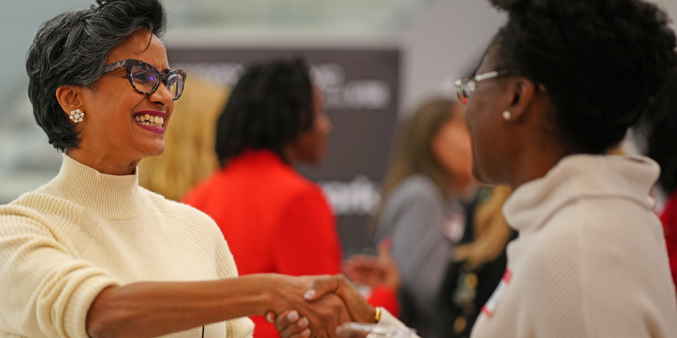 Two women smiling and shaking hands at an indoor event. One wears glasses and a white turtleneck, the other wears a cream sweater, with a blurred group of people in the background.