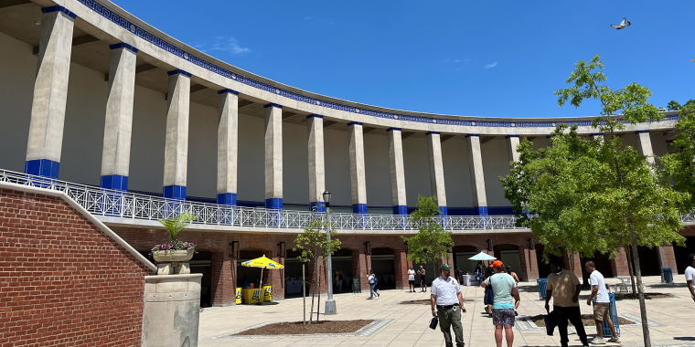 People walk in a sunny open plaza with large trees inside a curved building featuring tall columns and blue accents. A few yellow umbrellas provide shade, and a bird is flying in the clear blue sky.