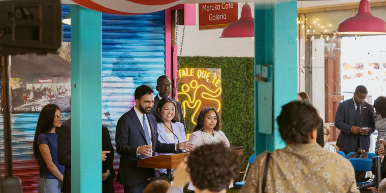 Mayor Mamdani and a group of people stand at a podium giving a speech in the colorful indoor space of La Marqueta with hanging lights and a crowd watching. The atmosphere is lively and festive, with blue and pink decor and a sign reading “Maruka Café Galería."