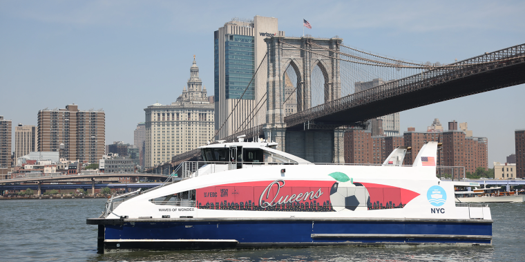 A white and blue NYC ferry with Queens and a soccer ball design sails on the river, with the Brooklyn Bridge and New York City skyline visible in the background.