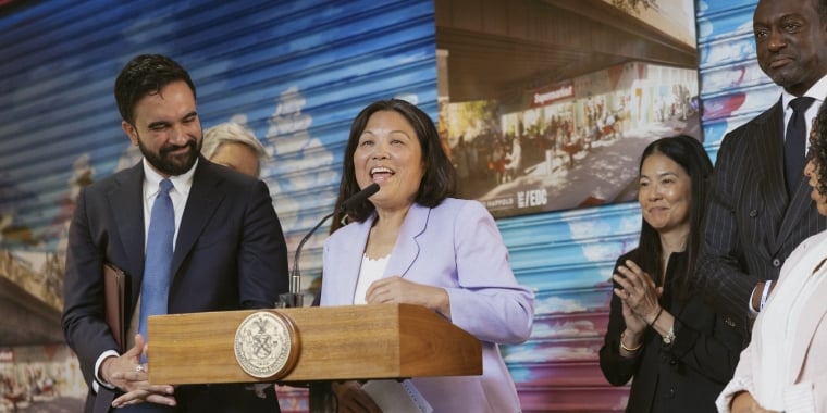 Deputy Mayor Julie Su in a light purple blazer speaks at a podium with a New York City seal, smiling, surrounded by Mayor Mamdani and NYCEDC members, in front of a colorful mural and a poster of a city street scene. Image credit: Kara McCurdy, Mayoral Photography Office.