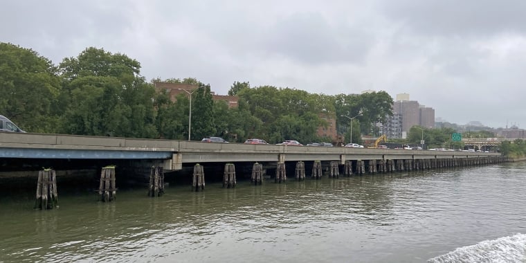 A gray, cloudy day over a riverside highway with cars driving along it. The highway is supported by wooden pilings, with trees and brick buildings visible in the background, and water in the foreground.