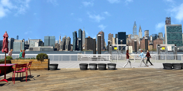 People walking and sitting on a wooden waterfront deck with planters and red tables; the New York City skyline and East River are visible under a clear blue sky in the background.