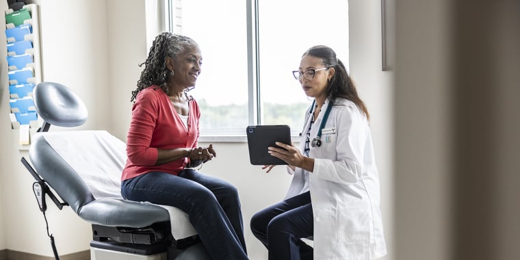 A doctor sits and shows a tablet to a patient who is seated on an exam table in a medical office. The patient smiles and listens as the doctor explains something, with natural light coming in through the window.