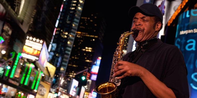 A man wearing a dark shirt and cap plays a saxophone at night on a busy, brightly lit street filled with colorful signs and billboards.