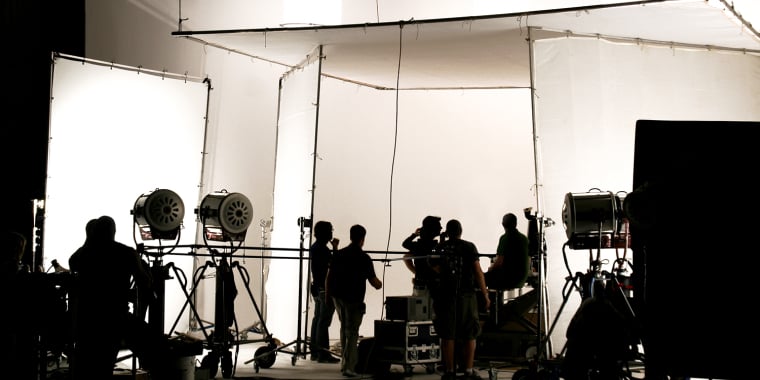 Silhouetted crew members work on a film set with large lights, cameras, and equipment against a white backdrop, creating a busy production atmosphere.