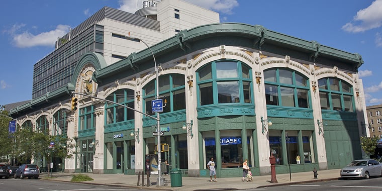Ornate, historic building with teal trim and large windows on a city street corner, housing a Chase bank. Modern additions are built on the roof. People walk on the sidewalk; traffic signals and street signs are visible.