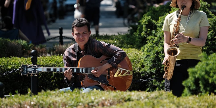 A man playing an electric guitar and a woman playing a saxophone perform together outdoors on a sunny day, surrounded by green bushes and people in the background.