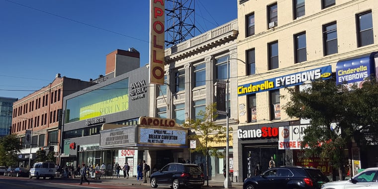 Street view of the Apollo Theater with its vertical red sign and marquee, surrounded by shops like Cinderella Eyebrows and GameStop, cars parked along the road, and a clear blue sky overhead.
