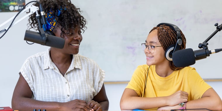 Two people sit at a table with microphones and audio equipment, smiling at each other as they record or broadcast. One wears headphones and a yellow shirt; the other has a headscarf and striped shirt. A whiteboard is in the background.