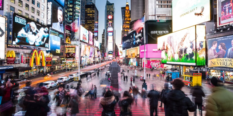 A bustling scene in Times Square, New York City, with blurred crowds, bright lights, colorful billboards, and advertisements illuminating the area at dusk.