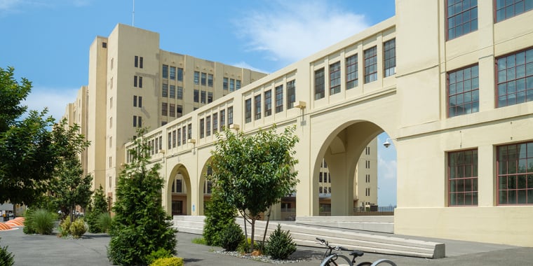 A large, light-colored building with arched walkways and many windows stands under a blue sky. In the foreground, there are trees, a bicycle parked at a rack, and a paved walkway.