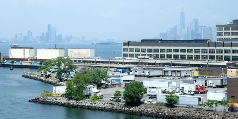 A view of a waterfront industrial area with warehouses, trucks, and greenery; a body of water is in the foreground, and a hazy city skyline is visible in the distance.