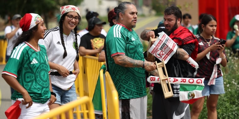 A group of soccer fans in green Mexico jerseys stand behind a yellow barrier, smiling and holding flags. One man holds a wooden noisemaker, and others wear festive hats and accessories. It is a festive outdoor setting.
