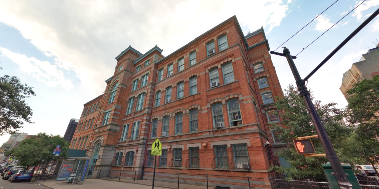 Streetview image of Julia de Burgos Latino Cultural Center showing a large, red-brick, multi-story school building with tall windows and a distinctive corner tower, set along a Harlem street with a sidewalk, street signs, and a few pedestrians in the foreground.