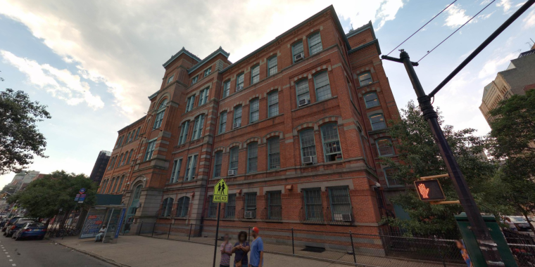 Streetview image of Julia de Burgos Latino Cultural Center showing a large, red-brick, multi-story school building with tall windows and a distinctive corner tower, set along a Harlem street with a sidewalk, street signs, and a few pedestrians in the foreground.