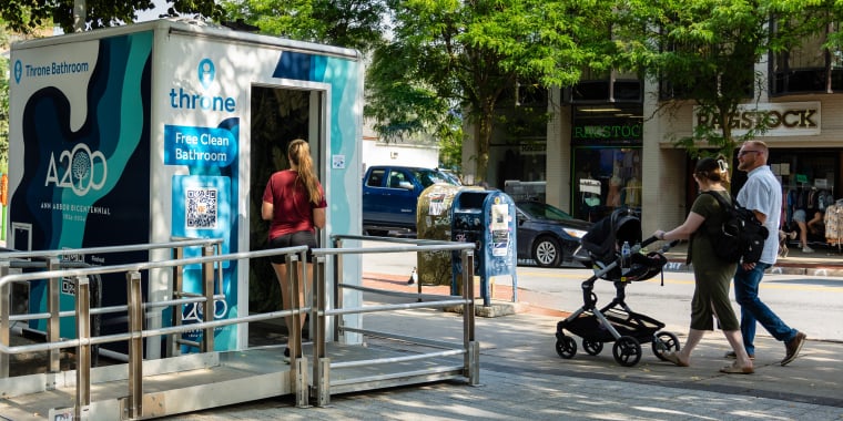 A woman waits to use a Throne public bathroom unit on a New York City sidewalk while a couple with a stroller walks by. Trees, parked bikes, and storefronts are visible in the background. Photo by Kem Davis.