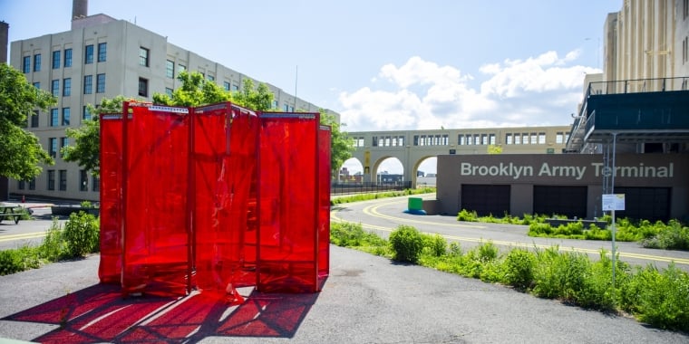 A large outdoor art installation made of translucent red panels stands in front of the Brooklyn Army Terminal, with a sunny sky, greenery, and industrial buildings in the background.