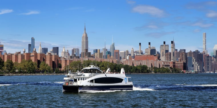 A New York City ferry travels on the East River with the Manhattan skyline, including the Empire State Building, visible in the background on a clear, sunny day.