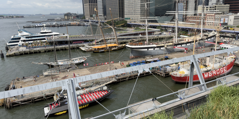 Several historic and modern boats docked at a New York City waterfront, with tall buildings in the background and people walking along the piers.