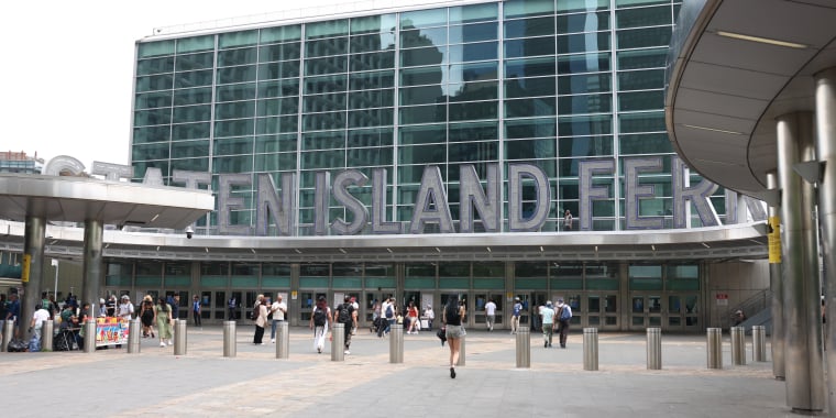 People walk in front of the Staten Island Ferry terminal, a large glass building with “Staten Island Ferry” in big silver letters above the entrance. Some street vendors are set up near the front.