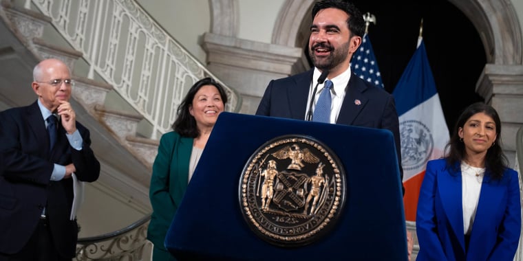 Mayor Mamdani speaks at a podium with the New York City seal, flanked by three people smiling. A staircase and NYC and American flags are in the background.