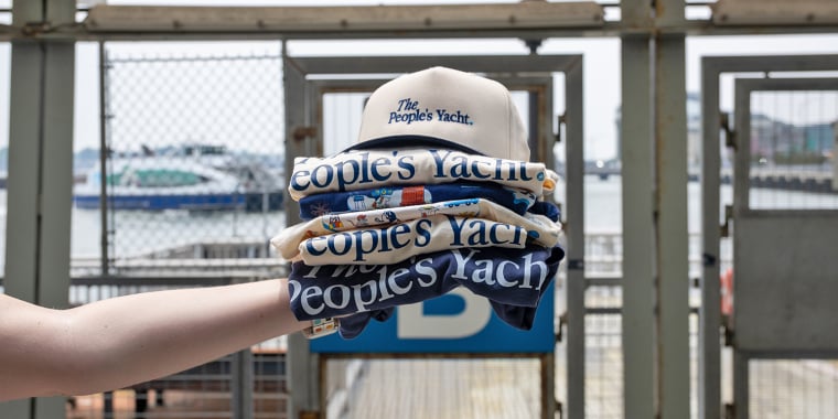 A hand holds a stack of folded The Peoples Yacht shirts and a matching hat at a waterfront dock, with boats and a city skyline visible in the background.