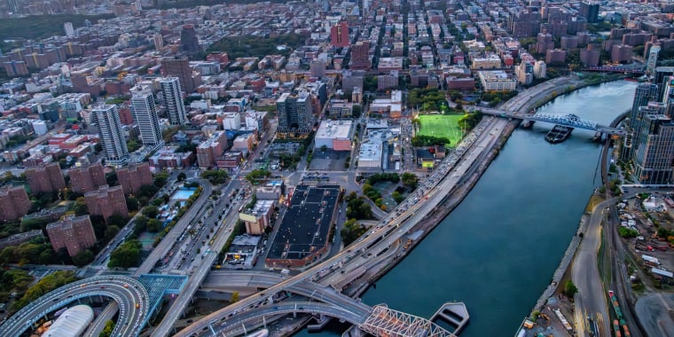 Aerial view of New York City at sunset, showing a river, several bridges, high-rise buildings, busy roads, and a park with green fields.