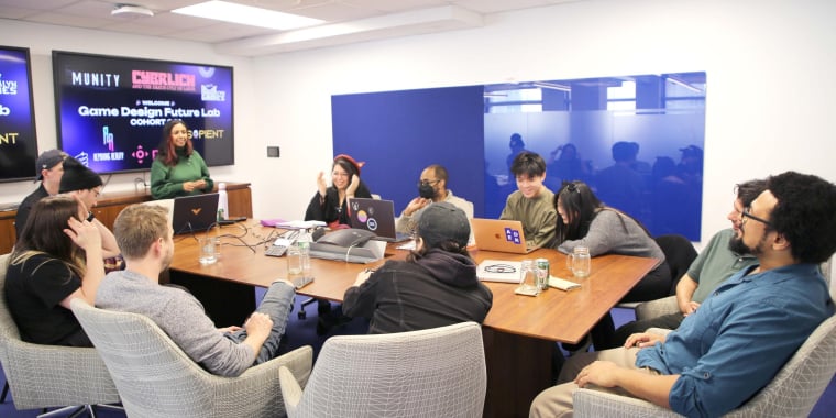 A diverse group of people comprising the first cohort of Game Design Future Lab, sit around a conference table, listening to Mitu Khandaker standing at the front. Laptops and drinks are on the table. Credit: NYU Tandon School of Engineering.