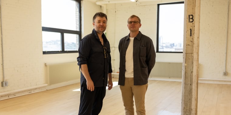 Stickbulb Co-Founders Russell Greenberg and Christopher Beardsley stand in their newly leased space at Building A, MADE Bush Terminal. The room is bright, with wooden floors, large windows, and white walls. Both men are wearing casual clothing and glasses. Sunlight is streaming in, and part of a pillar marked B1 is visible.