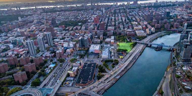 Aerial view of New York City at sunset, showing a river, several bridges, high-rise buildings, busy roads, and a park with green fields. The sky is orange and blue as the sun sets in the distance.