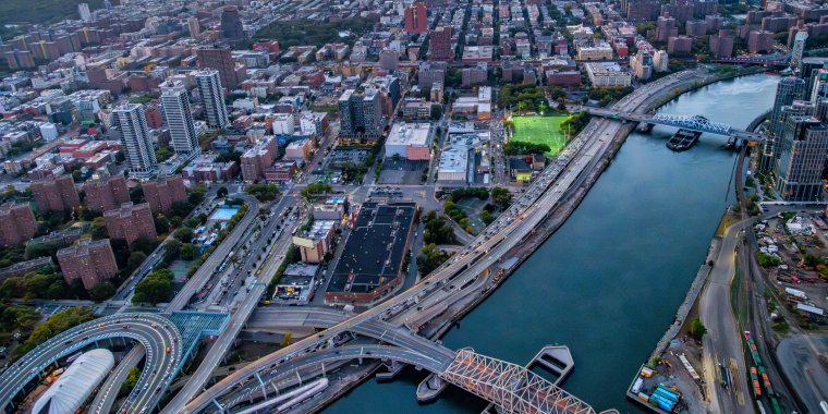Aerial view of New York City at sunset, showing a river, several bridges, high-rise buildings, busy roads, and a park with green fields. The sky is orange and blue as the sun sets in the distance.