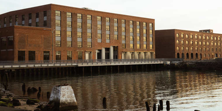 Photos of buildings A and C at MADE Bush Temrinal shows large, rectangular brick buildings with many windows beside a calm waterfront, reflecting in the water. Nearby, weathered wooden posts emerge from the shoreline. The scene is peaceful and bathed in soft light.