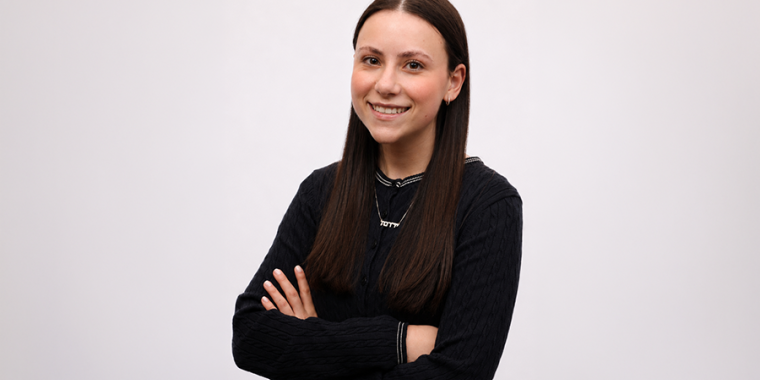 Jordana Stern in long brown hair smiles while standing with her arms crossed. She wears a black sweater and a necklace, and is posed against a plain white background.