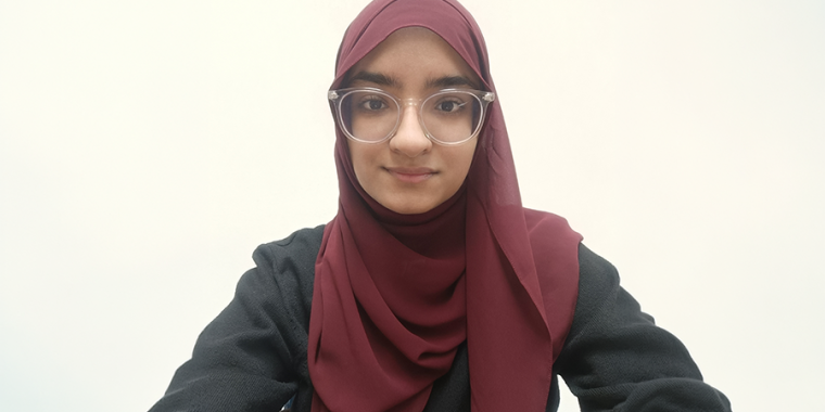 Maryam Ilyas in glasses and a maroon hijab is facing the camera and smiling slightly. The background is plain and light-colored.