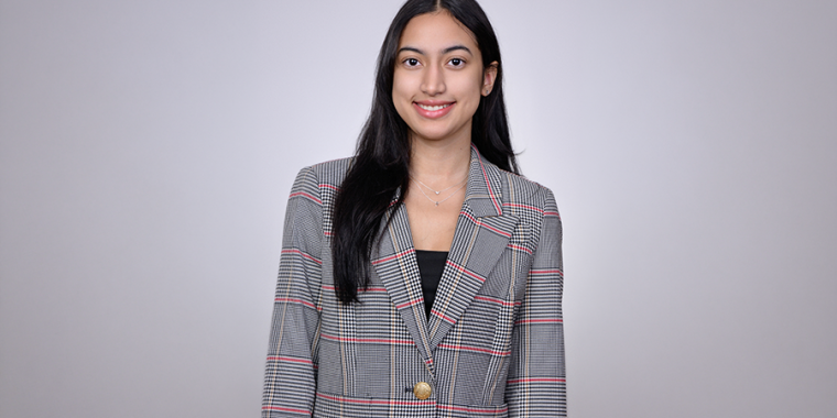 Pamela Rivera in long dark hair smiles, wearing a plaid blazer with a gold button over a black top, standing against a plain light gray background.